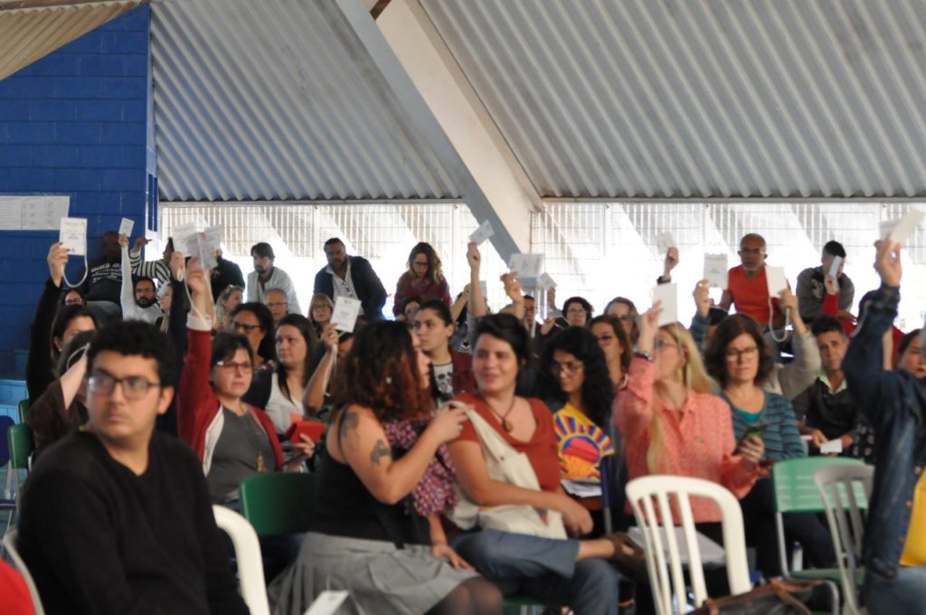 Imagem de um grupo de pessoas sentadas em cadeiras, sob um teto branco. Elas compareceram à primeira conferência de avaliação do Plano Municipal de Educação, em Campinas, São Paulo.