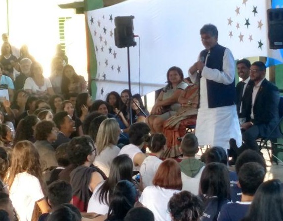 Fotografia da visita do indiano Kailash Satyarthi, vencedor do prêmio Nobel da Paz 2014, ao Centro de Ensino Médio Urso Branco (CEMUB). Ele discursa em pé, em frente a um toldo branco, e estudantes acompanham sentados no pátio da escola.
