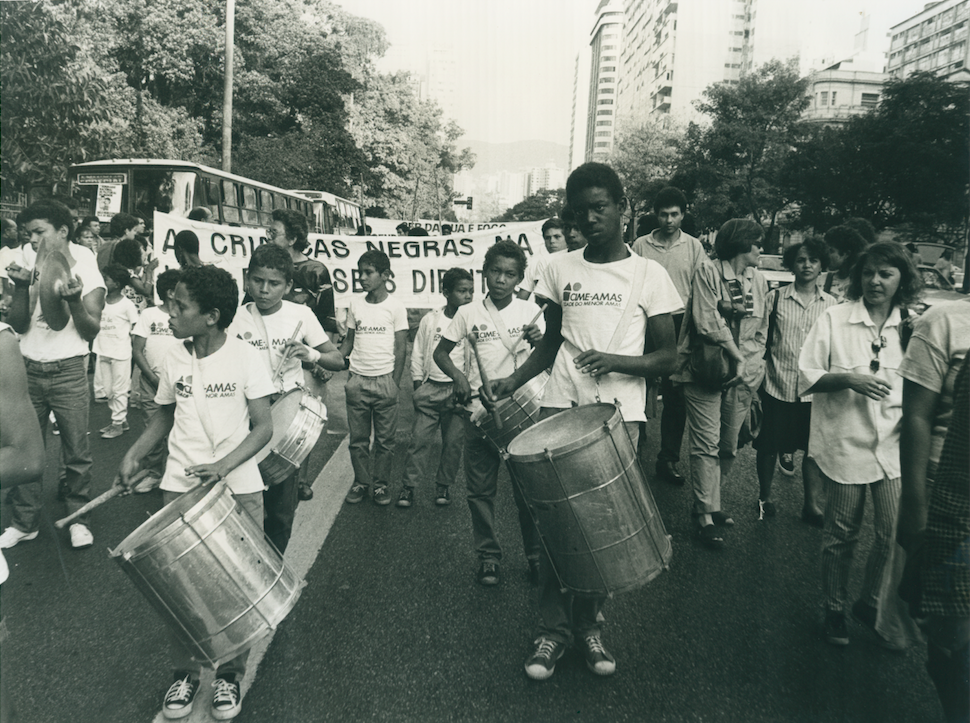 Imagem de manifestação de crianças e adolescentes pelos seus direitos. À frente das pessoas que compõem a manifestação, crianças e adolescentes tocam instrumentos de percussão. 